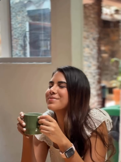 Una mujer con los ojos cerrados disfruta un café en taza verde junto a una ventana de la casa.