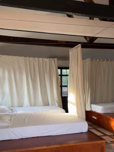 Dormitorio compartido con camas de plataforma en madera, colchones blancos y cortinas color crema bajo vigas de madera a la vista.