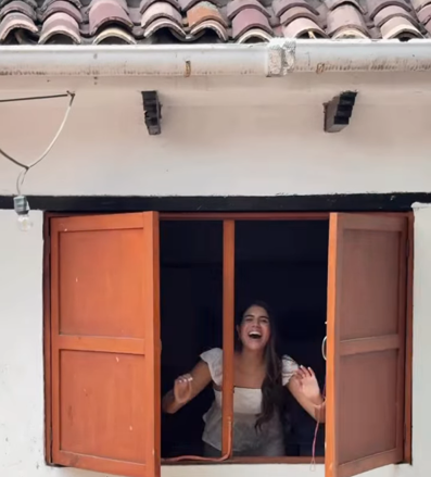 Fachada blanca con tejado de barro y postigos de madera naranja abiertos; una mujer se asoma riendo por la ventana.