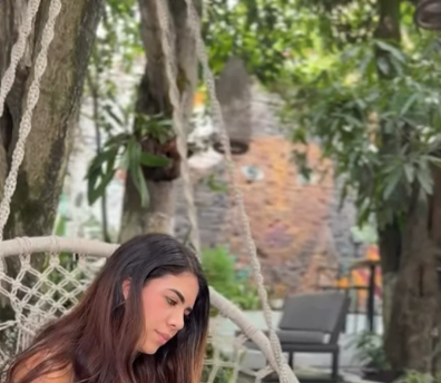 Una mujer lee sentada en una silla colgante tejida, bajo los árboles y junto a un muro de ladrillo.