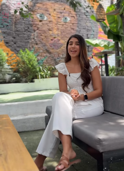 Una mujer sentada en un sofá gris de la terraza, frente a un mural colorido pintado sobre ladrillo, con plantas y una lámpara de mimbre.