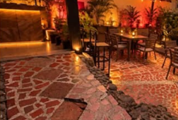 El patio de noche: piso de ladrillo y piedra, mesas y plantas bajo luces rojas y ámbar.