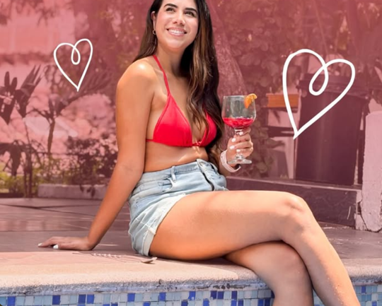 Una mujer sentada en el borde de la piscina de baldosas azules sostiene un coctel con rodaja de naranja; la foto original trae dos corazones dibujados.