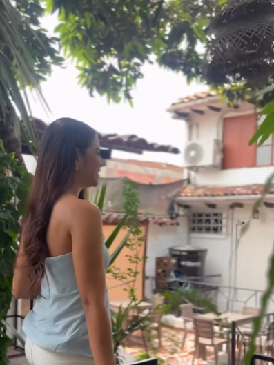 Una mujer mira hacia el patio entre plantas: mesas y sillas de madera, tejados de barro y una ventana con postigo naranja.
